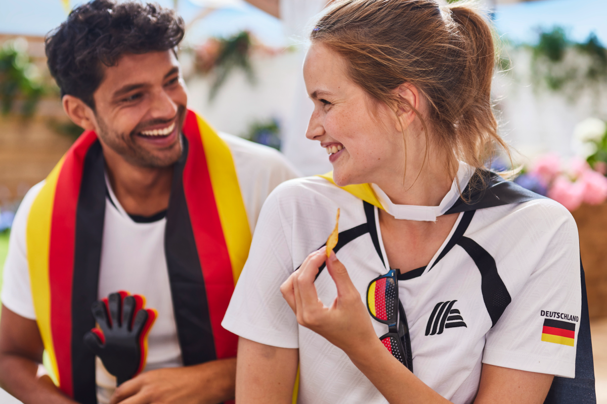 
Ein Mann und eine Frau im EM-Look der ALDImania-Kollektion lächeln sich auf der Fußball-Party im Garten an.